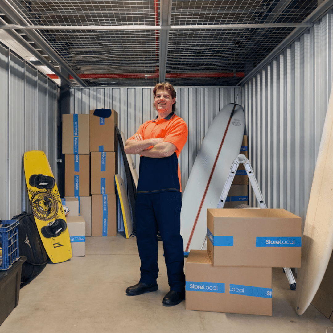 http://Tradie%20standing%20in%20storage%20unit%20with%20personal%20belongings%20like%20a%20surfboard%20and%20boxes%20being%20stored.