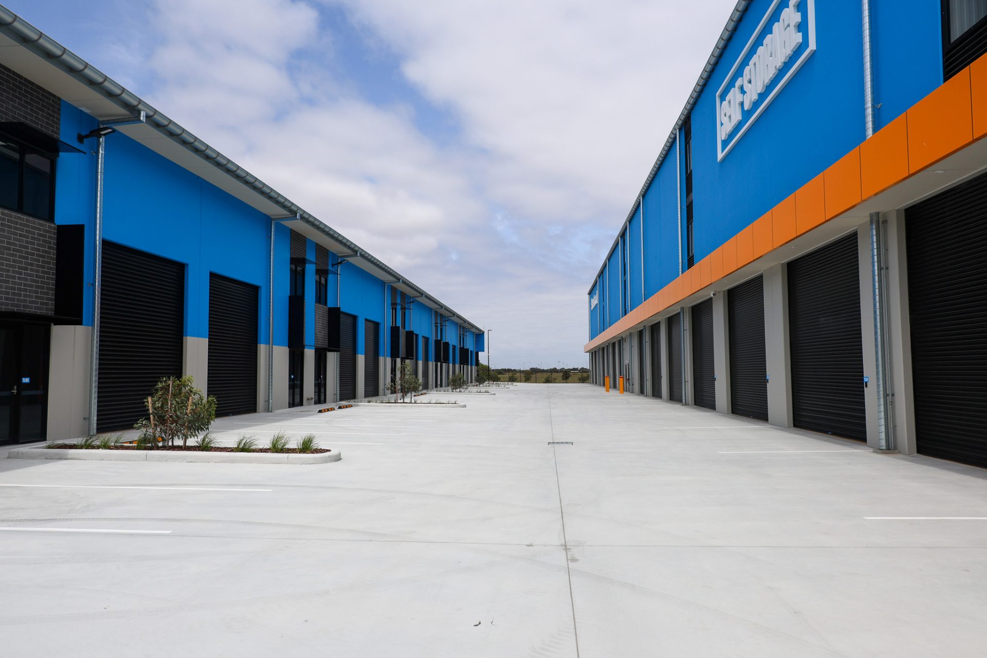 350A4555 A wide driveway between two rows of modern industrial storage buildings, featuring blue and gray facades with large black roller doors. A sign reading 'Self Storage' is displayed on the right building, and sparse landscaping is visible along the driveway.