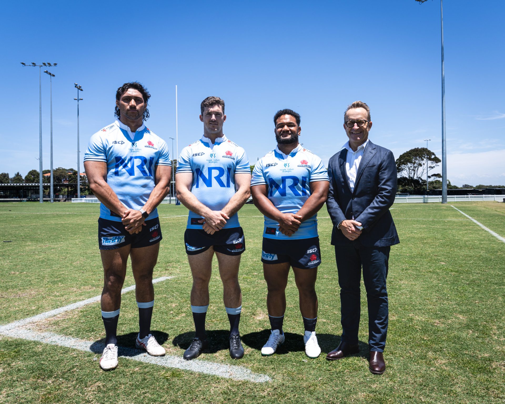 Store Local Announcement-1 Four individuals standing on a rugby field, including three NSW Waratahs rugby players wearing light blue jerseys with 'NRI' sponsor logos, black shorts, and socks. The players stand with their hands clasped in front, alongside a man in a dark suit on the right, likely a team official or sponsor representative. The scene is set under a clear blue sky, with rugby goalposts, green grass, and a few trees visible in the background